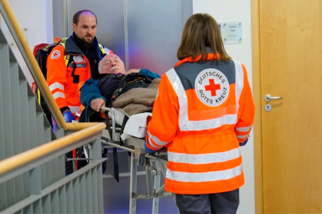 Zwei Sanitäter schieben einen Patienten auf einer Trage durch ein Treppenhaus