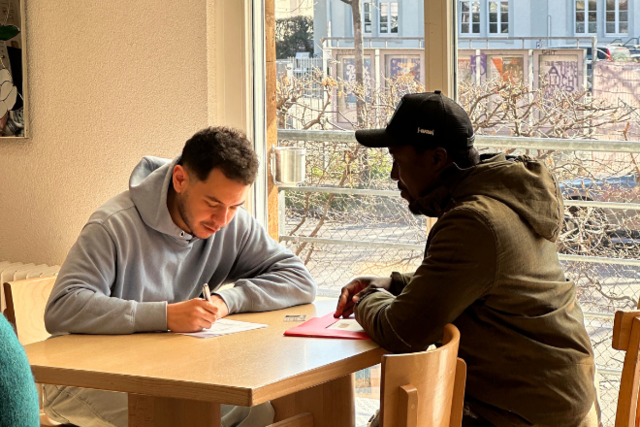 zwei junge Männer sitzen am Tisch, der eine hilft dem Anderen beim Ausfüllen eines Formulars