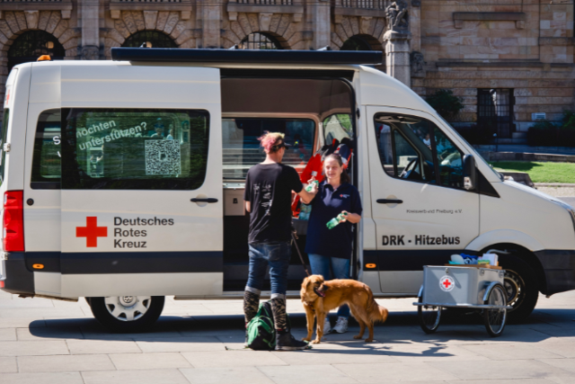 Der DRK Hitzebus steht in Freiburg, eine Mitarbeiterin reicht einem Mann eine Flasche Wasser