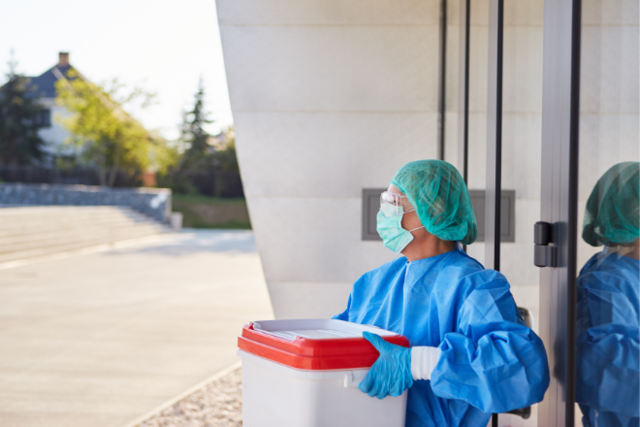 eine Frau in medizinischer Schutzausrüstung steht mit einer Kühlbox in der Hand vor einem Krankenhaus