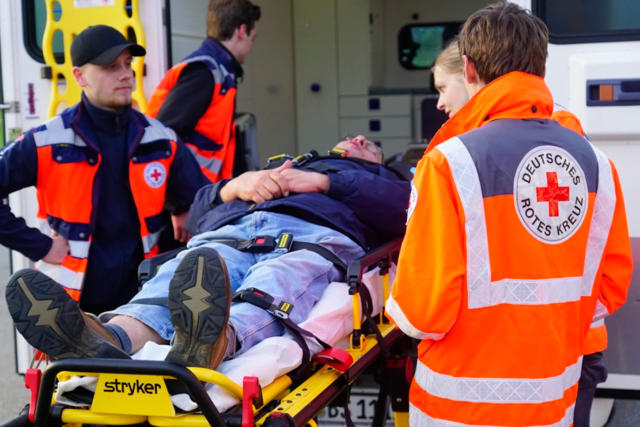 drei Helfer stehen in Einsatzkleidung um eine Trage, auf der ein Patient liegt