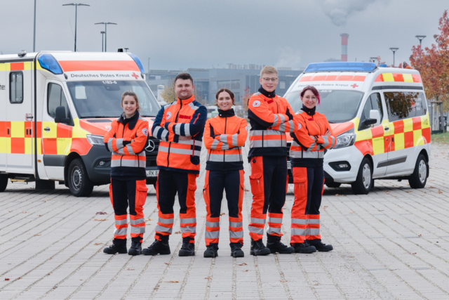 fünf junge Notfallsanitäter:innen stehen vor einem Rettungswagen und einem Krankenwagen und lächeln in die Kamera
