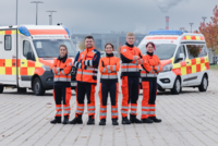 fünf junge Notfallsanitäter:innen stehen vor einem Rettungswagen und einem Krankenwagen und lächeln in die Kamera