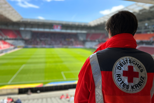 ein ehrenamtlicher Helfer in Einsatzjacke steht mit dem Rücken zur Kamera und schaut auf die Spielfläche des Freiburger Fußballstadions