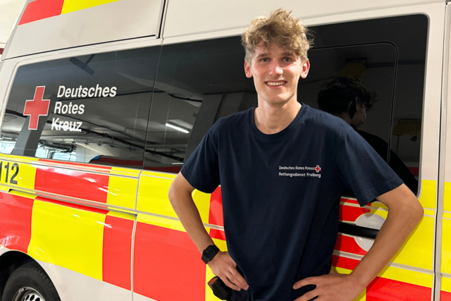 ein junger Mann steht in Dienstkleidung vor einem Krankenwagen und lächelt in die Kamera