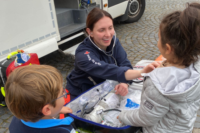 eine junge Notfallsanitäterin zeigt zwei Kindern lächelnd wie ein Stethoskop funktioniert