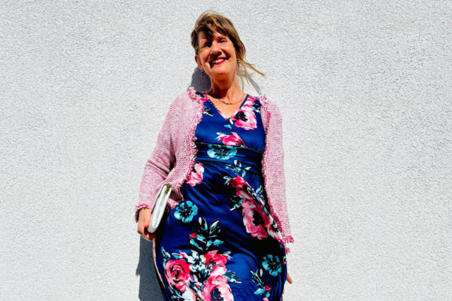 eine Frau steht in einem bunten Kleid vor einer Wand und lächelt in die Kamera